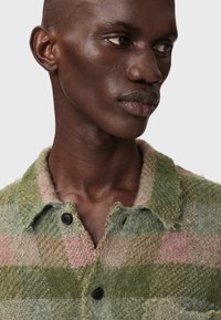 Πλεκτό πουλόβερ με πράσινο και ροζ καρό σχέδιο, γιακά, μαύρα κουμπιά και ακανόνιστη υφή, που τονίζει το μαλακό υλικό και τον σχεδιασμό με στρώσεις.