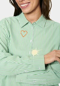 Camicia a righe verde e bianca con una grafica del sole giallo e un ricamo a forma di cuore arancione sul lato sinistro del petto. Design con bottoni.