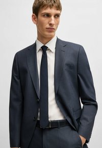 Giacca da completo a scacchi blu navy con revers a punta, due bottoni e tasca. Abbinata a una camicia bianca e una cravatta scura.