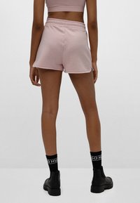 Světlé růžové bavlněné šortky s elastickým pasem, které mají volný střih a hladkou texturu, v kombinaci s černými kotníkovými ponožkami a botami.