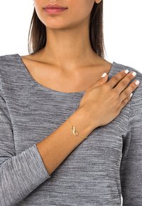 Guldarmband med en design av oändlighetssymbol, tillverkat av metall med en delikat kedja. Modellen har målade naglar och bär en grå topp.