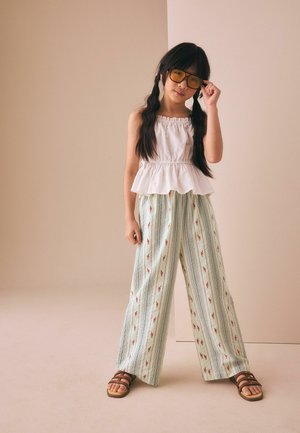 Mädchen mit langen dunklen Zöpfen trägt gelb getönte Brille, weißes ärmelloses Oberteil, weite gestreifte Blumenhose und braune Sandalen, stehend.