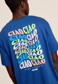 Blauwe katoenen t-shirt met meerdere kleuren "CIAO" tekst in speels gerangschikte lettertypen op de achterkant, met "BUON VIAGGIO" eronder gedrukt.