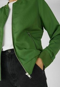Groene bomberjack met een gladde textuur, voorzien van een ritssluiting aan de voorkant, geribbelde manchetten en een zijzak, gecombineerd met een wit shirt en zwarte broek.