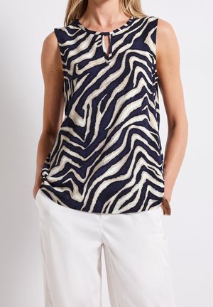 Vrouw draagt een mouwloze marineblauwe en witte zebraprint blouse met een kleine keyhole aan de voorkant, gecombineerd met witte broek en een houten armband.