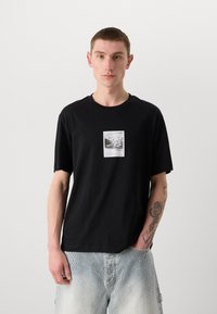 T-shirt in cotone nero con collo rotondo e maniche corte, caratterizzato da una stampa grafica centrale in bianco e nero. Vestibilità casual.