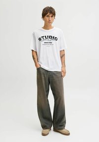 Hvid oversized T-shirt med sort grafisk tekst, parret med løse, falmede denimjeans og lyse tan slip-on sko. Neutral baggrund.
