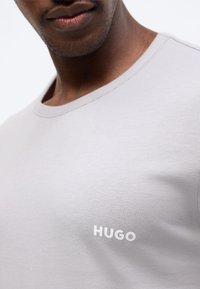 Κοντινό πλάνο ενός ατόμου με σκούρο χρώμα δέρματος που φοράει ανοιχτό γκρι μπλουζάκι με το λευκό λογότυπο «HUGO» στο αριστερό στήθος.