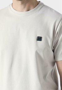 T-shirt di cotone grigio chiaro con scollo rotondo e maniche corte, caratterizzato da un piccolo logo quadrato nero sulla parte superiore sinistra del petto.