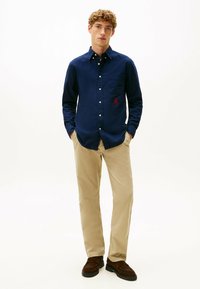 Navy button-up shirt met een borstzak en een rood logo, gecombineerd met beige broeken en bruine instapschoenen. Gladde stof, klassieke pasvorm.