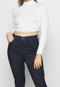 Un crop top blanc à col roulé côtelé associé à un jean en denim sombre à taille haute. Le haut présente des manches longues et un design ajusté.