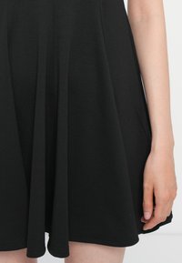 Robe sans manches noire avec une jupe évasée, en tissu lisse. Elle présente des plis verticaux et un col rond, avec des ourlets réguliers.