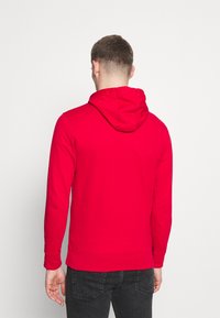 Felpa rossa con cappuccio regolabile, maniche lunghe e polsini a costine. Il tessuto sembra liscio e leggermente elastico, aderente in vita.