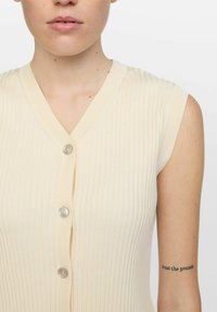 Ljusbeige ärmlös stickad topp med ribbad textur, med en V-ringning och tre knappar på framsidan.