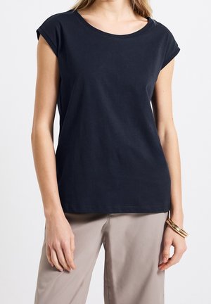 Vrouw die een eenvoudig zwart shirt met korte mouwen en lichtbeige broek draagt, met gouden armbanden aan haar rechterpols.