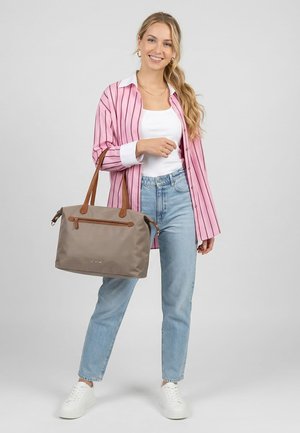 Lächelnde Frau in rosa gestreiftem Hemd, weißem Oberteil, blauen Jeans, weißen Sneakern, die eine taupefarbene Handtasche mit braunen Griffen und Reißverschluss hält.