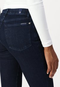 Jean de mezclilla oscura con un diseño elegante y una cintura alta, costuras sutiles y una pequeña etiqueta en el bolsillo trasero. Textura suave, forma ajustada.