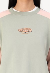 Žalias džemperis su rožinėmis pečių detalėmis, banguotu kaklu ir siuvinėtu "Billabong Australia" logotipu persikų spalvos. Minkšta medvilnė.
