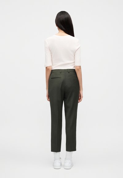 Γυναίκα με μακριά σκούρα μαλλιά στέκεται γυρισμένη πλάτη, φορώντας ένα ανοιχτό ροζ κοντομάνικο τοπ, σκούρο πράσινο παντελόνι κομμένο ελαφρώς, άσπρες κάλτσες και άσπρα παπούτσια.