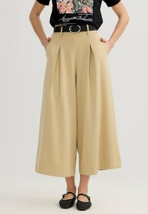 Mujer con pantalones cortos de pierna ancha beige con pliegues delanteros, cinturón negro, camiseta negra con estampado floral y zapatos Mary Jane negros tejidos.