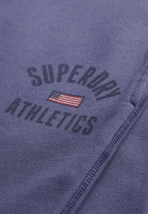 Paarse stof met de tekst "SUPERDRY ATHLETICS" en een kleine geborduurde Amerikaanse vlag tussen de woorden, zichtbare stiksels aan de zijkant.