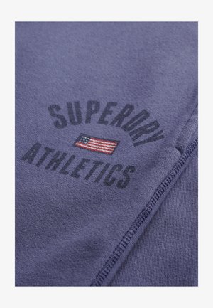 Lila Stoff mit dem Text "SUPERDRY ATHLETICS" und einer kleinen gestickten amerikanischen Flagge zwischen den Wörtern, sichtbare Nähte entlang der Seite.