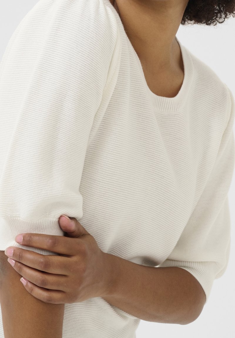 Femme portant un haut blanc texturé à manches courtes, tenant son bras gauche avec sa main droite sur un fond clair uni.