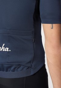 Tamnoplava atletska majica kratkih rukava s džepom na patentni zatvarač i bijelim tekstualnim detaljem. Glatka, prozračna tkanina s prilagođenim dizajnom.