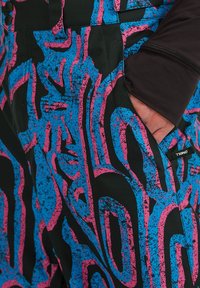 Zwarte broek met een gedurfde abstracte print in blauw en roze, met een gladde textuur en een detail van een zijzak. Merkenlabel zichtbaar.