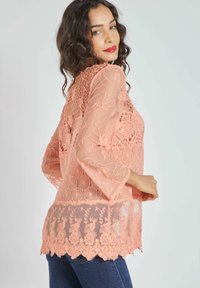 Blusa de encaje melocotón con un patrón floral, fabricada en tejido transparente, con mangas acampanadas, desviación en el bajo y detalles de bordado intrincado.