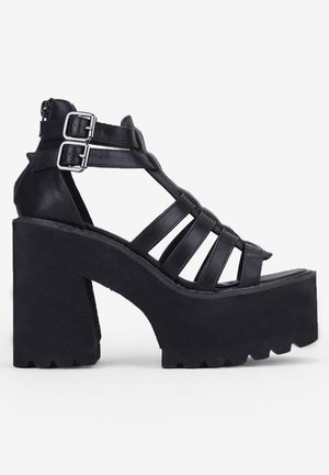 Sandalias negras de tacón alto de cuero con suelas de plataforma gruesas y texturizadas, que cuentan con múltiples correas y doble hebilla para el cierre.