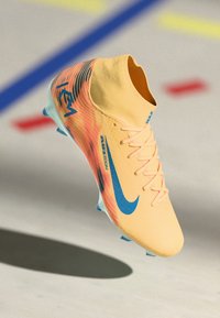 Fußballschuh in Gelb mit blauer Nike-Swoosh und "Air Zoom"-Text, schwebend über dem Hallenfußboden mit farbigen Linien.