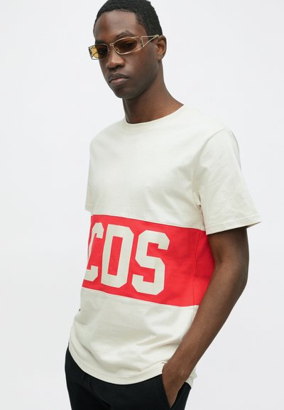 T-shirt a maniche corte in cotone bianco sporco con una banda orizzontale rossa che presenta la scritta "COS" in bianco. Girocollo e vestibilità rilassata.