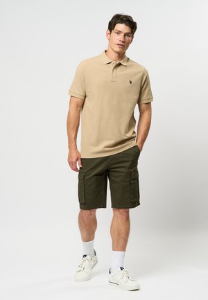 Beige polo shirt med tre-knap lukning og broderet logo, parret med grøn cargo shorts. Hvide sneakers og korte sokker fuldender outfittet.