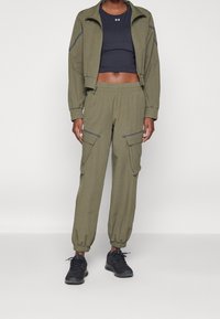 Olivgrön sportjacka med dragkedja och elastiska mudd, matchad med tillhörande avsmalnande cargobyxor med sidfickor och grå detaljer. Svarta sneakers.