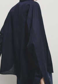 Chemise en lin marine avec une coupe ample, des manches larges et des fentes sur les côtés. Design minimaliste avec une texture douce et un léger éclat.