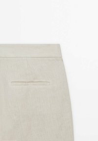 Gros plan d'un pantalon en lin beige avec une poche passepoilée horizontale à l'arrière et une ceinture plate sur fond blanc.