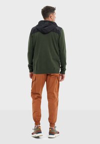 Un sweat à capuche vert avec une capuche noire, associé à un pantalon cargo orange doté de poches latérales et de poignets élastiques. Les chaussures sont multicolores avec des lacets.