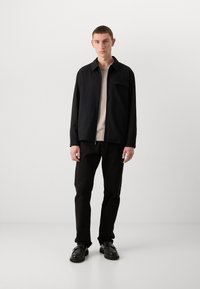 Black, textured jacket with a collar, front pocket; beige top underneath. Black pants and black shoes complete the outfit.