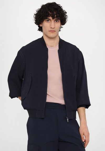 Mornarska bomber jakna s kratkim ovratnikom, zadrgo in elastičnimi manšetami. Obuta čez svetlo roza majico, z udobno prileganjem in detajli stranskih žepov.