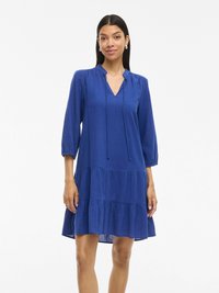 Plava haljina do koljena s rukavima do tri četvrtine, dekolteom u obliku slova V i detaljima s vezicom. Sadrži višestruke volane na rubu.