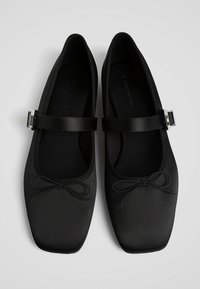 Une paire de ballerines noires de type Mary Jane avec des bouts carrés, de fins nœuds décoratifs à l'avant et des sangles à boucle en argent sur le dessus.