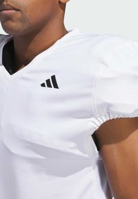 Jersey atlético blanco con diseño de cuello en V. Presenta el logo negro de Adidas en el hombro, tejido texturizado y mangas elásticas.
