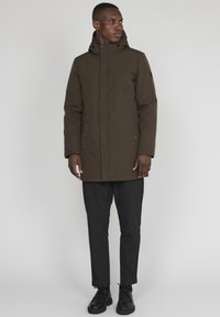 Jachetă parka impermeabilă, maro, cu glugă, închidere cu fermoar și capse, având un exterior texturat și un branding minimal. Purtați-o peste pantaloni negri.
