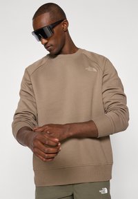 Bruine sweatshirt met geribde manchetten en zoom, voorzien van een klein logo in de linkerbovenhoek. Model draagt een paar zwarte oversized zonnebrillen.