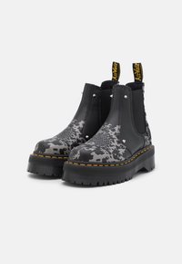 Black Chelsea boots with gray abstract pattern, thick textured sole, elastic side panels, and yellow stitching accents.