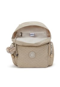 Sac à dos beige Kipling avec un compartiment principal ouvert, des poches zippées à l'avant et sur les côtés, et un porte-clés en forme de singe en peluche suspendu sur le côté gauche.