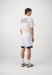 Camiseta blanca con estampado "AX" en la parte posterior, combinada con pantalones cortos blancos con un dobladillo negro. Diseño simple, corte relajado y estilo casual.