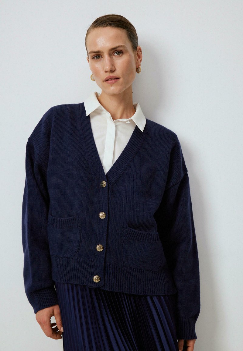 Námorně modrý pletený cardigan s výstřihem do V, předními kapsami a zlatými knoflíky. Textura je měkká a silueta je volná.