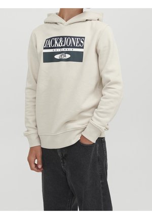 Person iført en beige Jack & Jones hættetrøje og sorte jeans, med den ene hånd delvist i lommen, mod en ensfarvet lys baggrund.